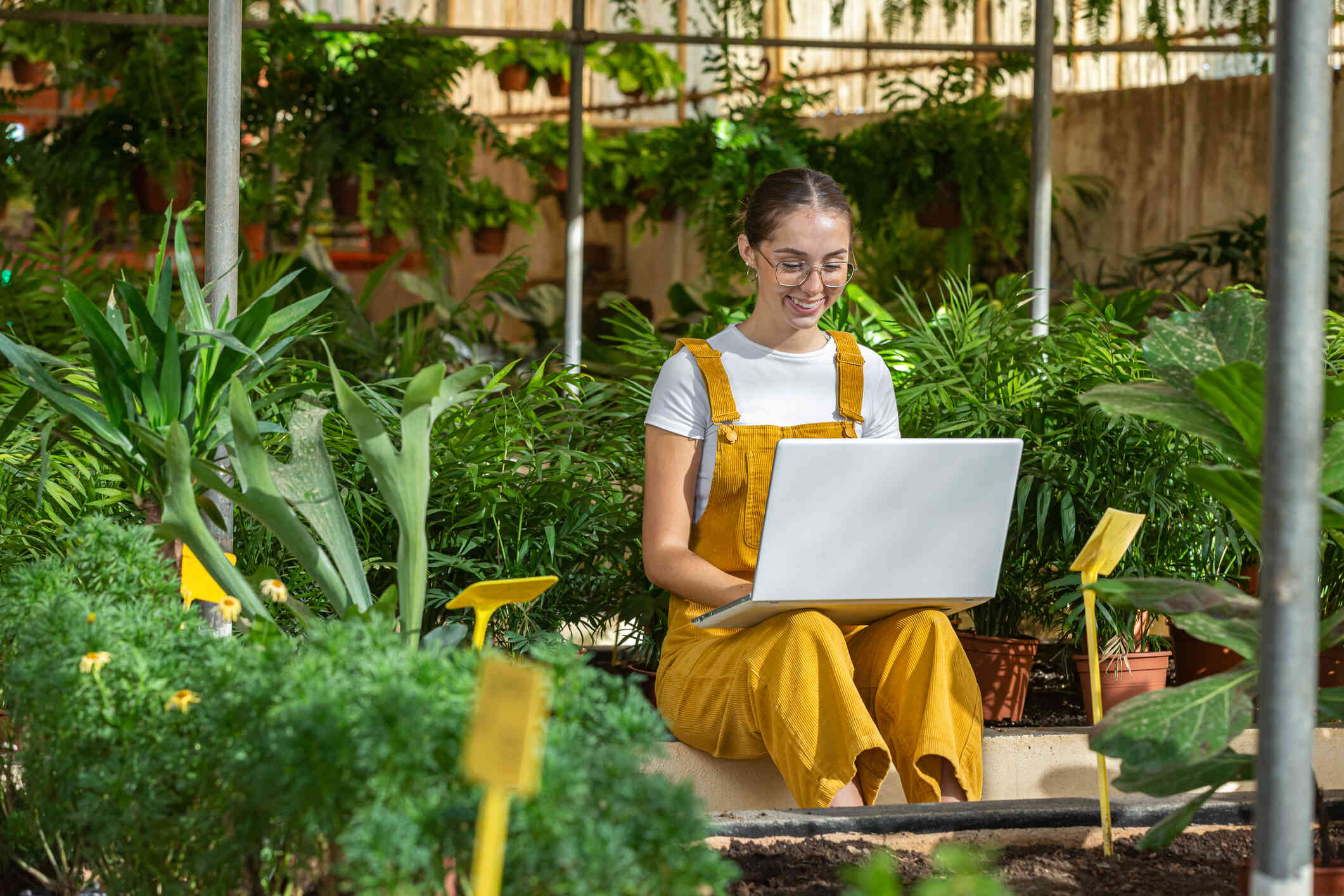 A woman in yellow overalls sits outside in a garden while smiling at her laptop