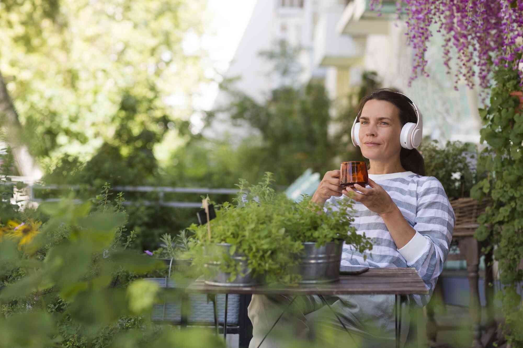 A woman sits outside on a patio with lots of greenery and wears over the ear headphones while holding a coffee mug.