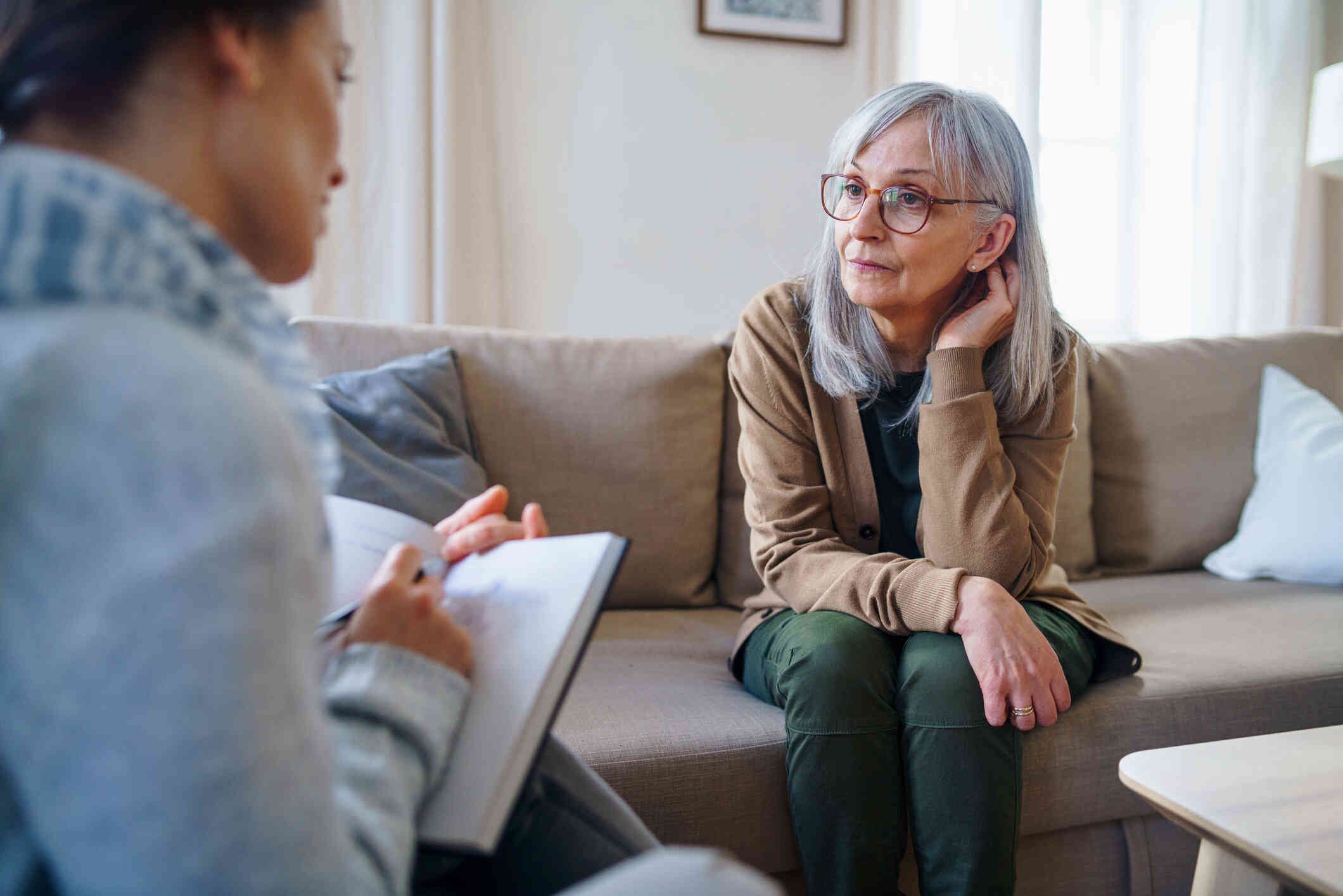 A mature woman wearing glasses has a serious expression as she sits on a couch across from another woman who is writing on a clipboard.