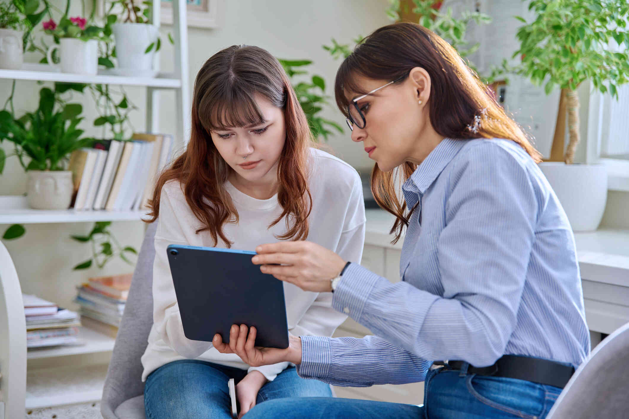 A woman and a young girl are seated together, both looking at the same tablet.