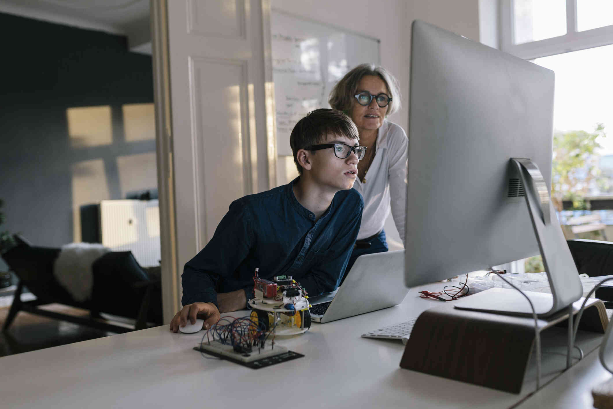 A young boy is seated at his desk, looking at the computer screen, while a woman stands behind him doing the same.