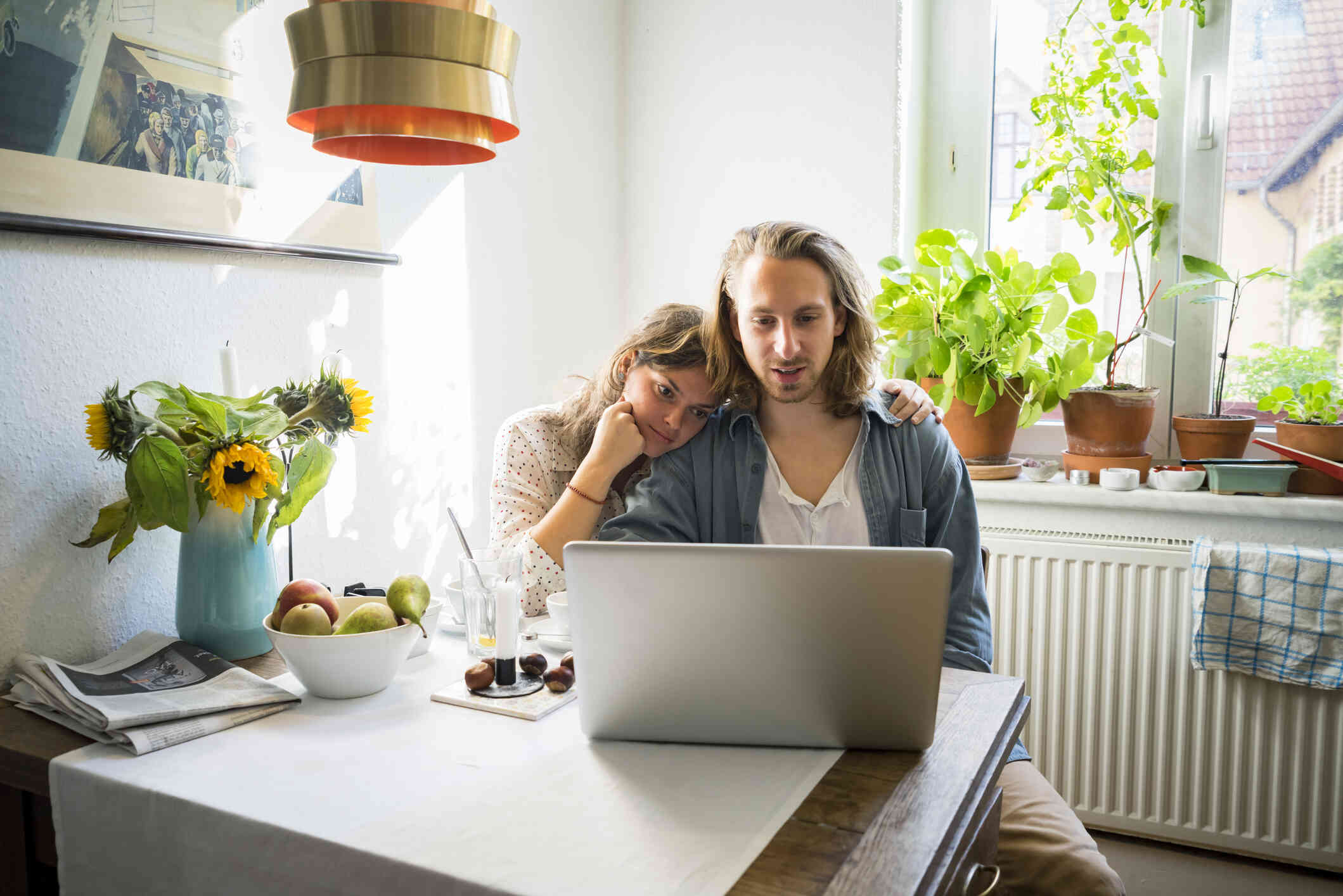 A couple sits at a dining table leaning on each other while looking at a computer