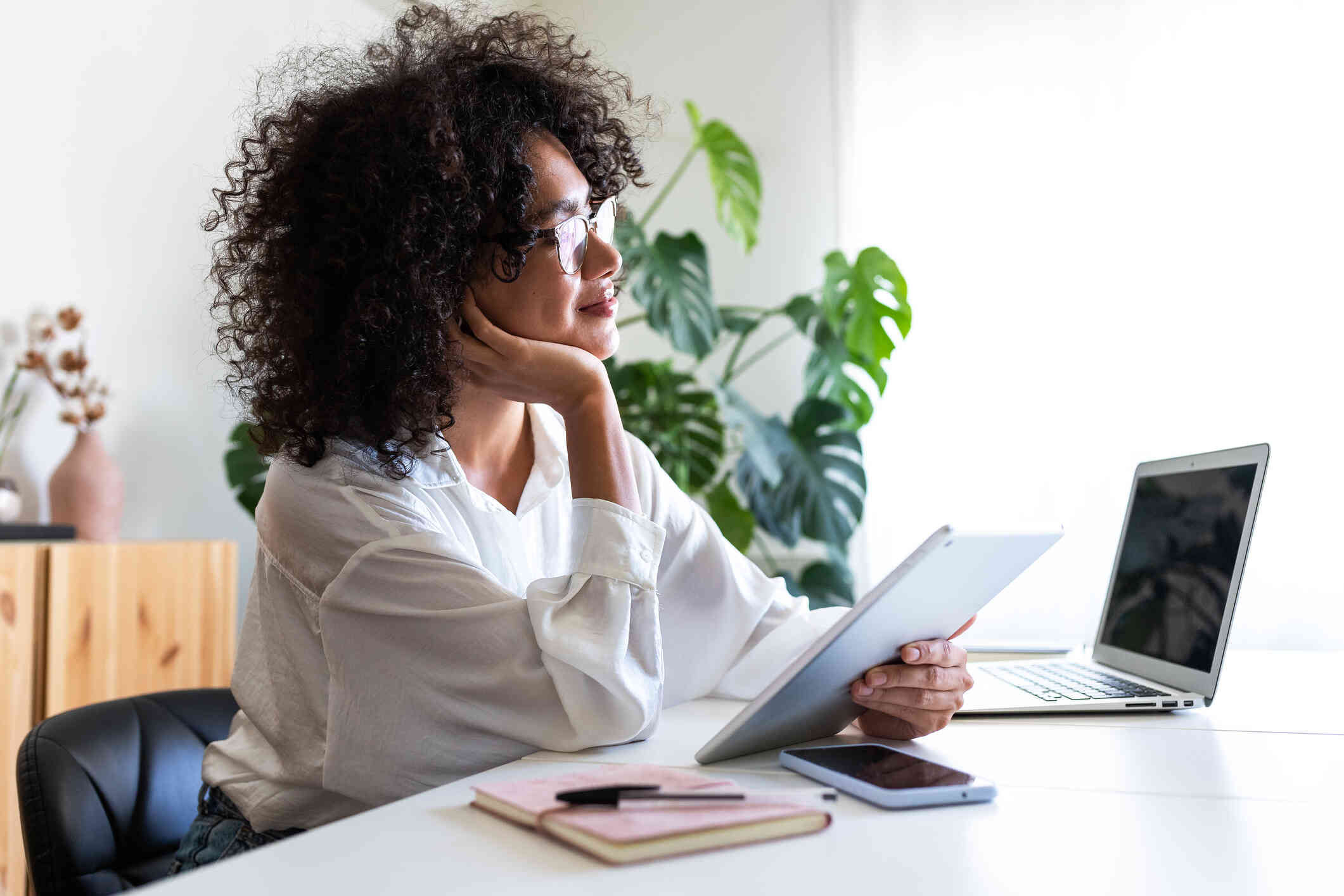 A woman in a white shirts smiles into the distance as she holds a tablet in her hand