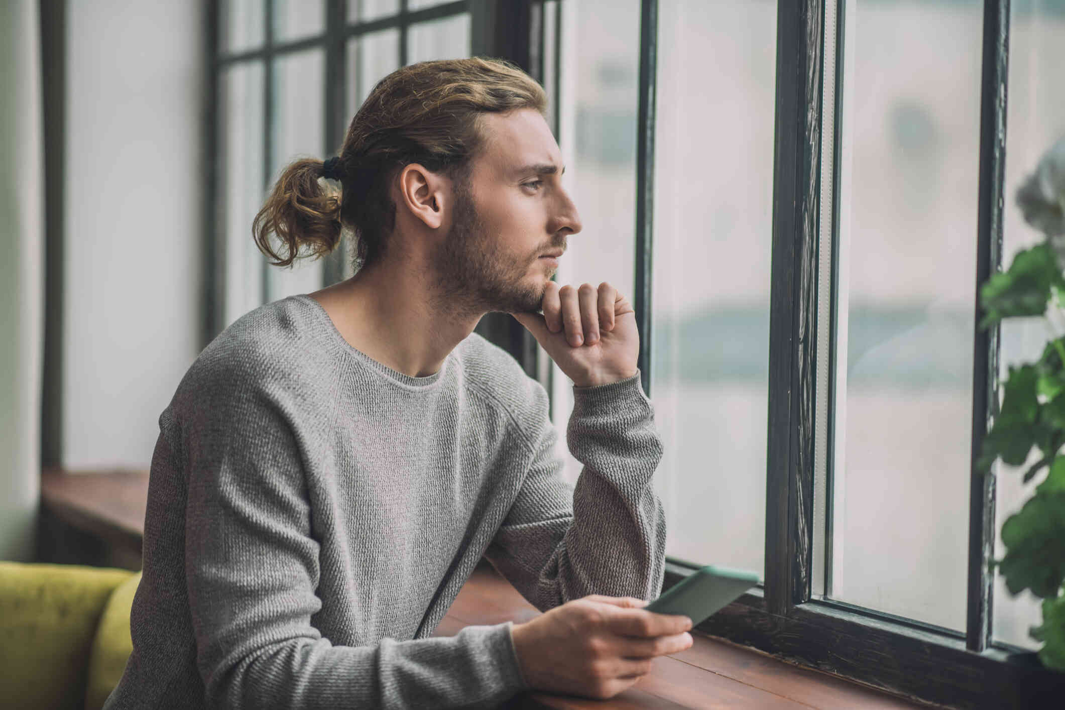A man with a ponytail is seated by the window, his hand touching his chin as he gazes outside pensively.